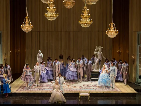 A theatrical production on stage features actors in elaborate period costumes, posing amidst chandeliers, statues, and a detailed set. The scene is well-lit and richly decorated.