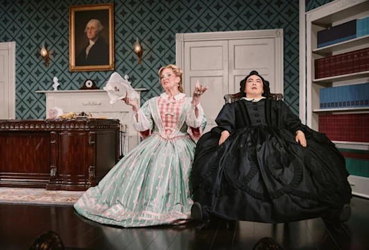 Two performers in period gowns seated in a drawing room, one in green plaid gesturing, the other in black.
