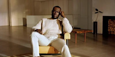 A man in a light-colored sweater and pants sits in a modern chair in a minimalist room with neutral tones and a log-filled fireplace.