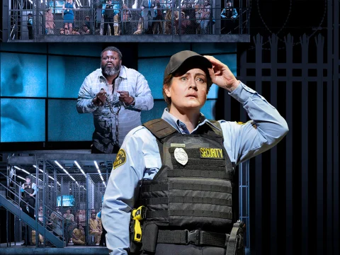 A security officer is in the foreground, adjusting their hat. In the background, a man in restraints stands in front of large screens, with a multi-level prison setting and inmates visible.