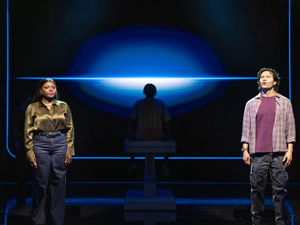 Three actors on a stage in front of a glowing blue screen, with one person seated on a pedestal between two standing actors.
