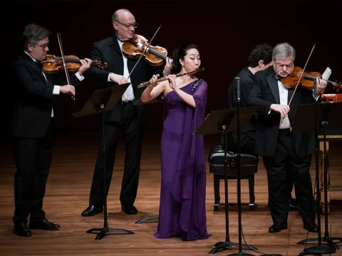 A group of five musicians performs on stage. Four play string instruments while one, dressed in a purple gown, plays the flute. Music stands with sheet music are positioned in front of each musician.