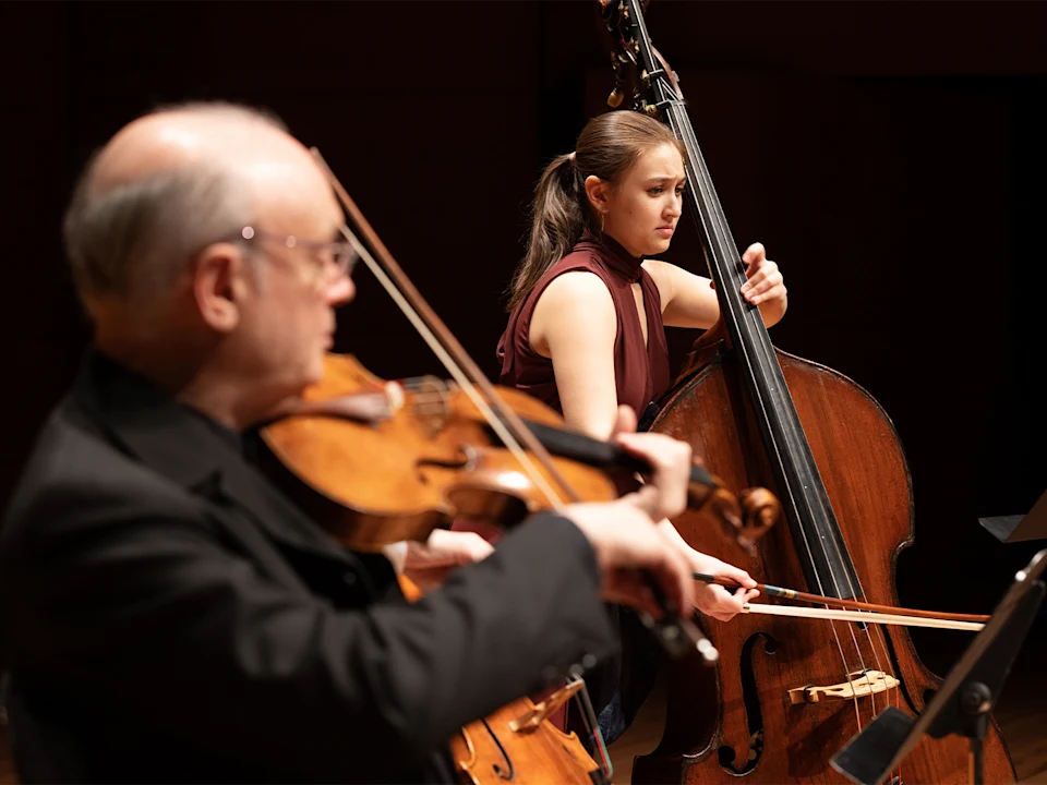 A young woman plays the double bass with a bow while an older man in the foreground plays the viola during a classical music performance.
