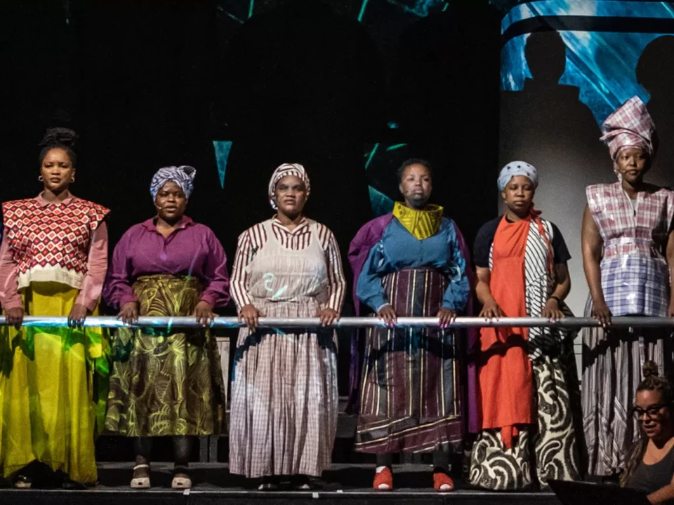 Six people stand side by side on stage, wearing a variety of colorful traditional and contemporary outfits with headwraps.