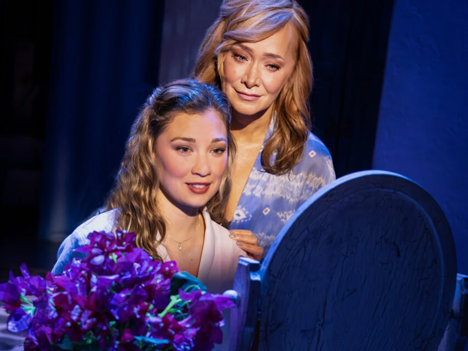 Two women standing in front of a mirror; one seated with curly hair and a white top, the other standing with wavy hair, wearing a patterned blouse. Purple flowers are in front of them.