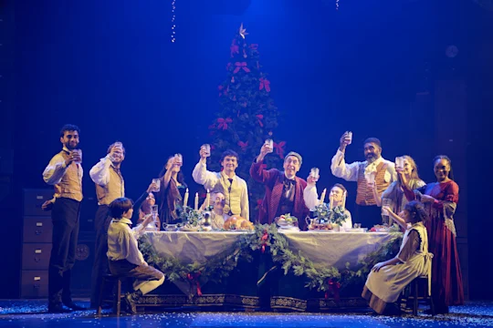 A group of people in period costumes sit and stand around a festive table, raising glasses in a toast, with a decorated Christmas tree in the background on stage.