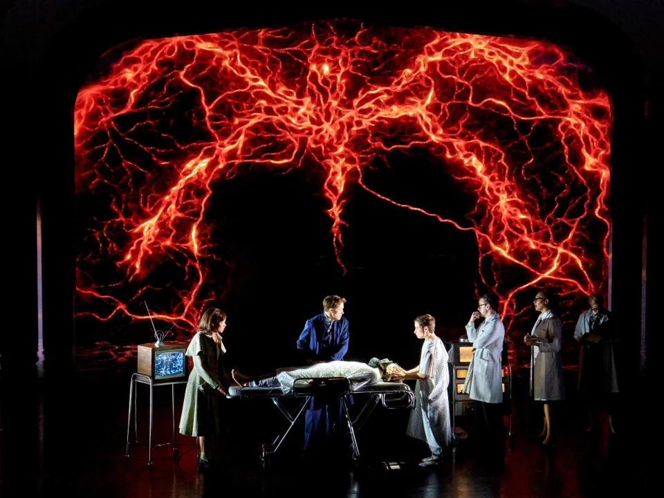 A theatrical scene depicts doctors and medical staff surrounding a patient on an operating table with a vivid red and orange electric backdrop.