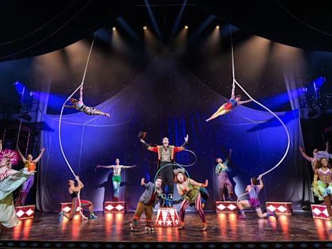 A circus-themed stage performance with acrobats on ropes, performers in colorful costumes, and a ringmaster at center stage under dramatic lighting.