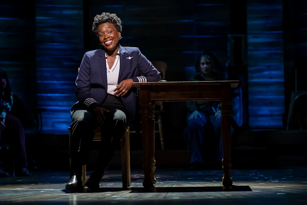 A person in a pilot’s uniform sits and smiles at a wooden desk on stage, with a blue-lit background and other people seated in the shadows.