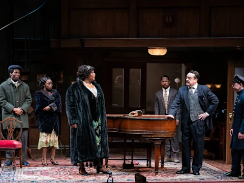 Six actors stand around a piano on a detailed stage set, dressed in 1920s-style clothing, suggesting a dramatic scene in a theater production.