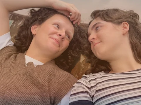 Two people with curly hair lie side by side, facing each other and smiling, one wearing a brown vest and the other a striped shirt.