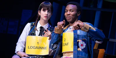 Two actors wearing numbered spelling bee contestant signs, "Logainne" and "Leaf," sit next to each other onstage, looking forward under stage lighting.