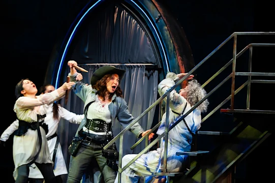 Three actors perform a dramatic scene onstage; one in a hat points a sword at another on stairs, while a third person restrains them, with an arched backdrop in the background.