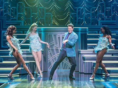 A man in a blue sequined jacket dances on stage between three women in matching sequined dresses.