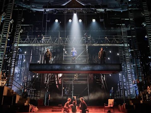 A theatrical stage set features actors standing on multi-level scaffolding with dramatic lighting highlighting the center figure; additional performers are gathered on the stage below.