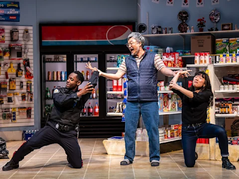 Three people are in a convenience store set; a man in a police uniform and a woman kneel dramatically on either side of an older man standing in the center with his arms raised.