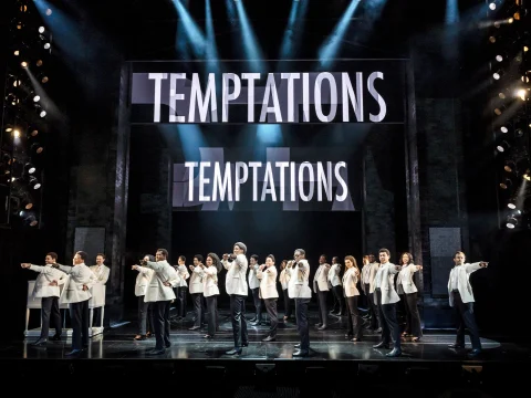 Performers in white jackets and black pants dance in formation on stage, with a backdrop displaying the word "Temptations" illuminated above them.