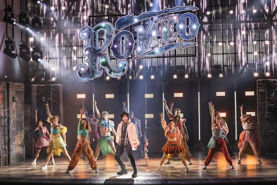 A group of performers dance on stage in colorful costumes with a large illuminated “Romeo” sign above them.