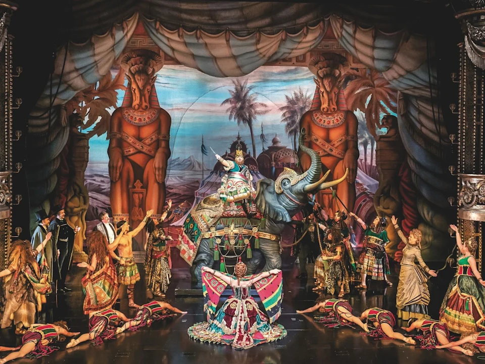 Actors in elaborate costumes perform a theatrical scene with an elephant prop and ornate backdrop featuring large statues, palm trees, and painted scenery.