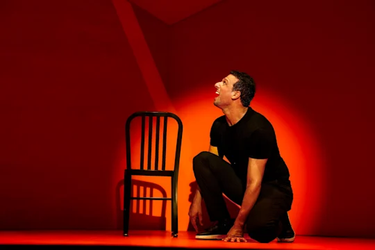 Production photo of Other in New York, showing a man in a black shirt kneels on the floor next to a black chair, dramatically lit by a red and orange spotlight on a minimalist stage.