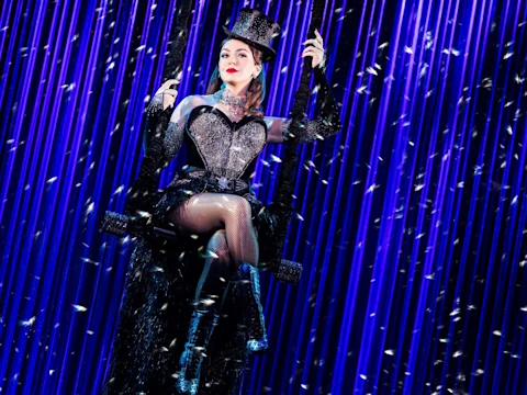 A performer in a sparkling black costume, top hat, and fishnet stockings sits on a swing against a backdrop of vertical blue lights and floating confetti.