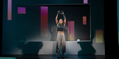 A person stands center stage with arms raised, in front of a geometric set with colored rectangular panels and dramatic lighting.