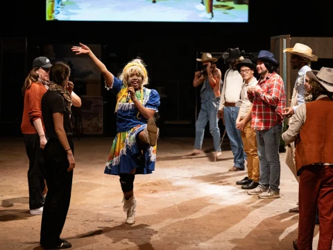 A person in a colorful dress stands on one leg and gestures enthusiastically in the middle of a semicircle of people, some in cowboy hats and casual attire, under dim lighting.