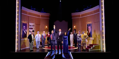 A group of actors stands on a brightly lit theater stage with gold walls, framed portraits, and spotlights, posing at the end of a performance.