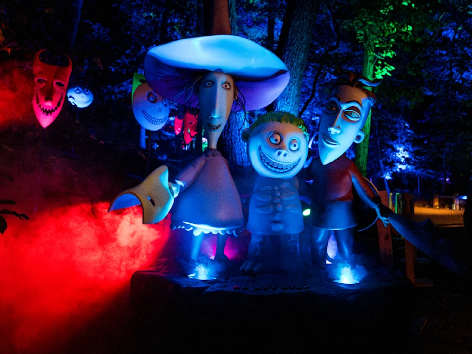 Four cartoonish, spooky figures with exaggerated features stand illuminated by colorful lights and surrounded by red fog in a nighttime forest setting.