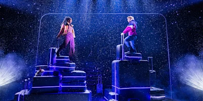 Two actors stand on stacks of suitcases on stage under artificial snow, with dramatic lighting and a dark background.