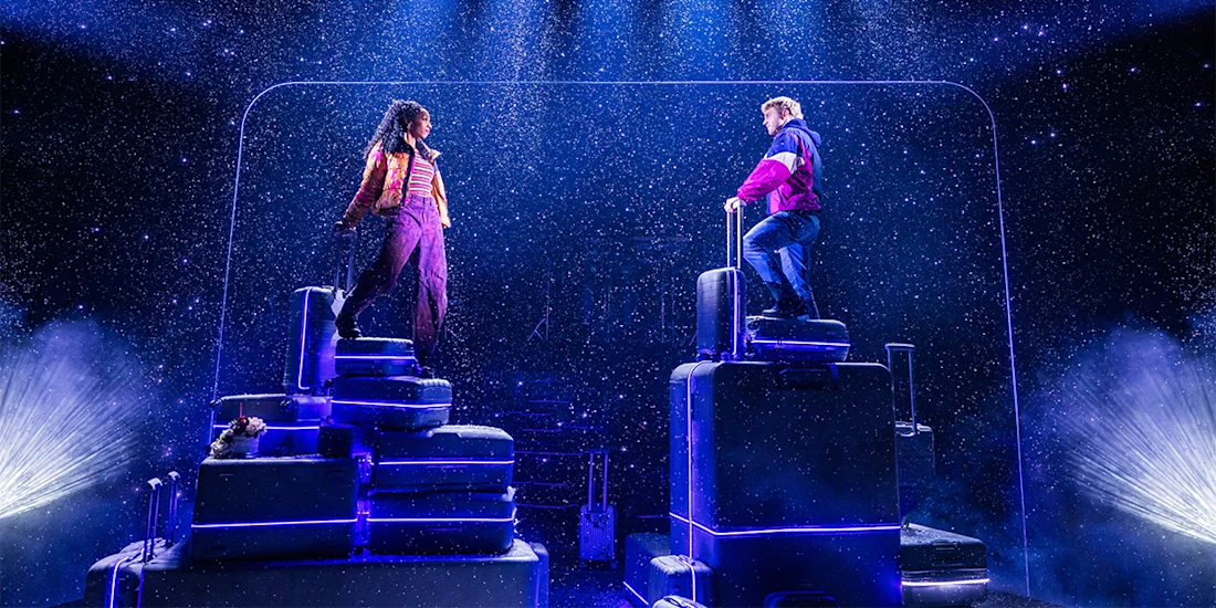 Two actors stand on stacks of suitcases on stage under artificial snow, with dramatic lighting and a dark background.
