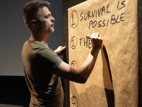 A person writes on a large paper with the words "Survival is Possible" at the top. They are standing in a dimly lit room.