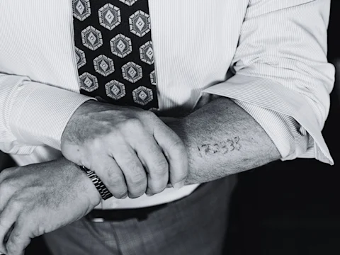 A person in a dress shirt and patterned tie reveals a faded number tattoo on their forearm.