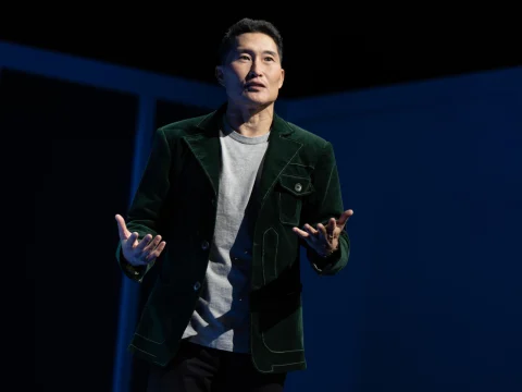 Person in a green jacket and gray shirt speaking on stage, gesturing with hands.