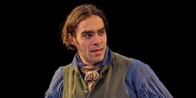 A person in period costume, featuring a blue shirt, green vest, and cravat, stands with hands on hips against a black background.
