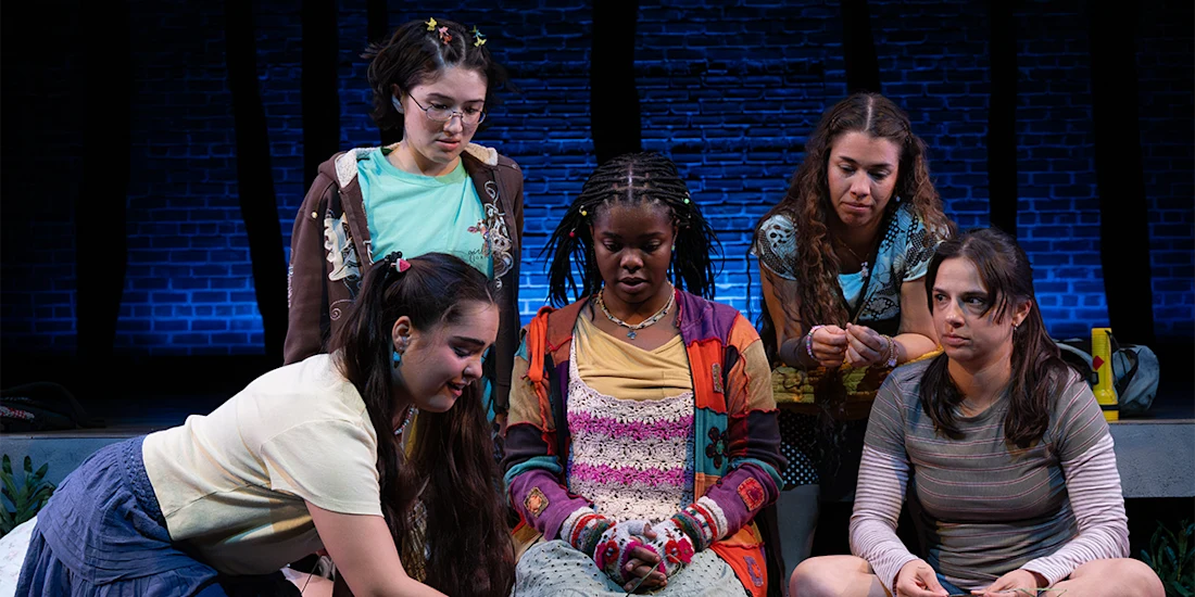 Five young women sit and kneel together onstage, dressed in casual, colorful clothing, with a brick wall and blue lighting in the background.