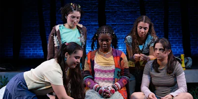 Five young women sit and kneel together onstage, dressed in casual, colorful clothing, with a brick wall and blue lighting in the background.