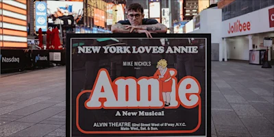 A person leans on a large display sign for “Annie: A New Musical” in Times Square, with colorful lights and buildings visible in the background.