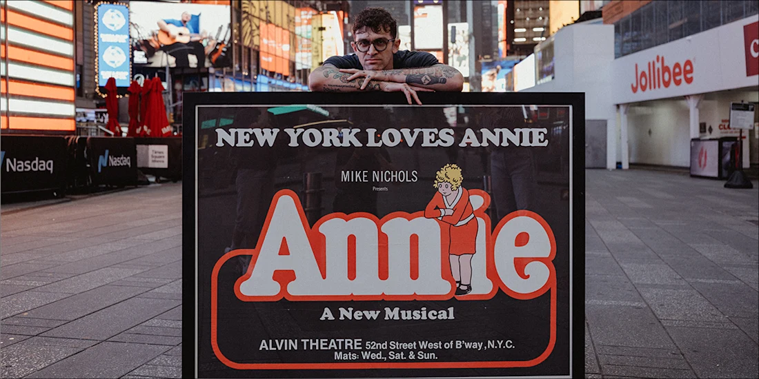 A person leans on a large display sign for “Annie: A New Musical” in Times Square, with colorful lights and buildings visible in the background.