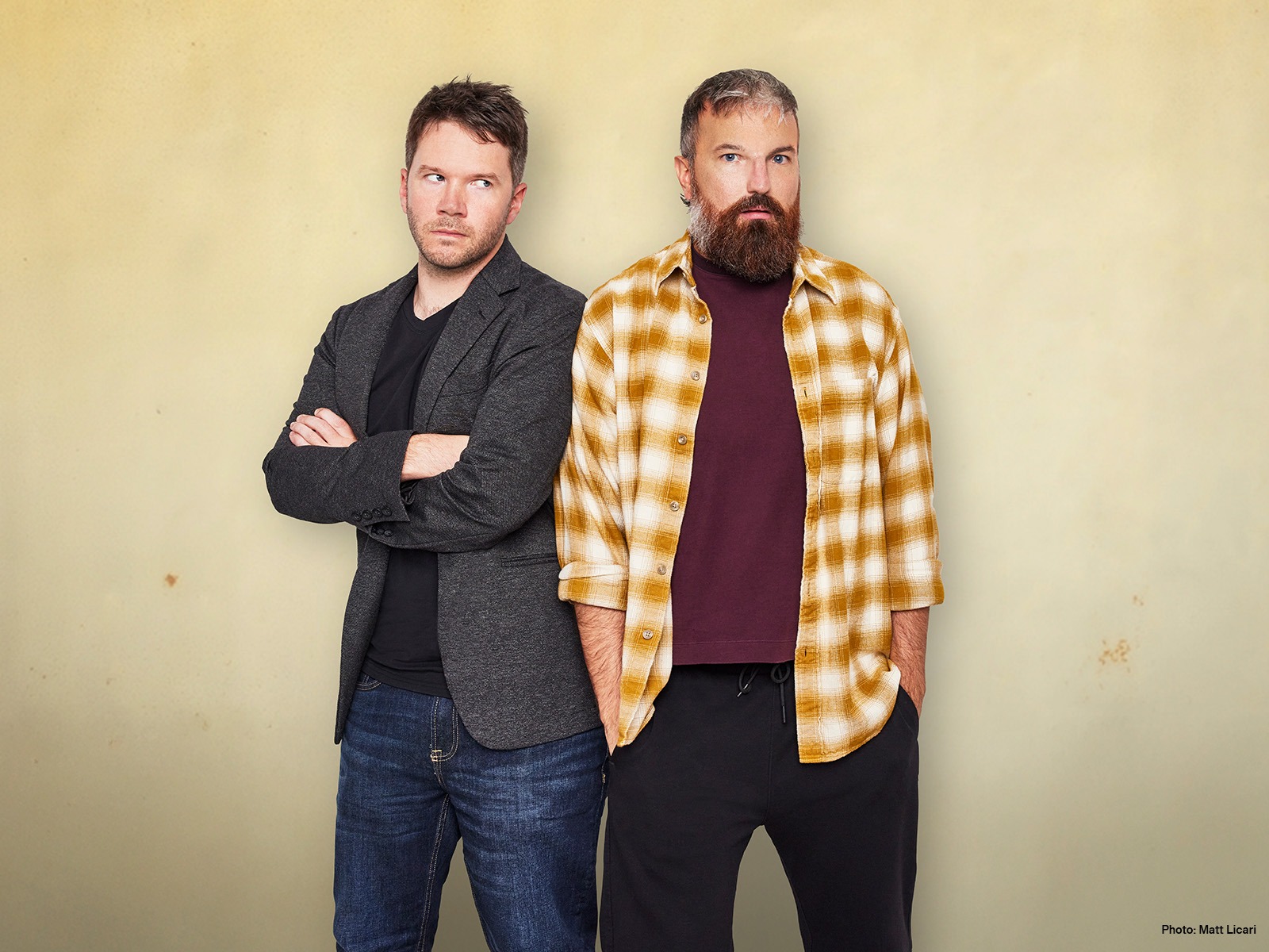 Two men stand in front of a beige wall; one has arms crossed and wears a gray blazer, the other wears a yellow plaid shirt over a maroon t-shirt and has hands in pockets.