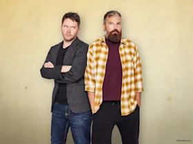 Two men stand in front of a beige wall; one has arms crossed and wears a gray blazer, the other wears a yellow plaid shirt over a maroon t-shirt and has hands in pockets.