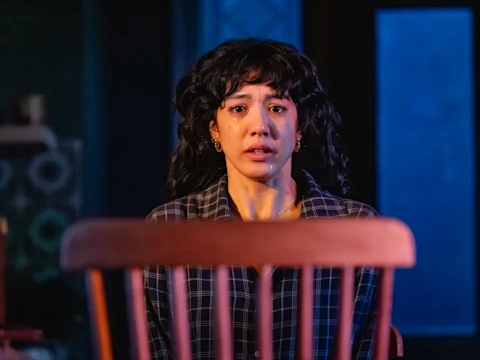 Production photo of Paranormal Activity in San Francisco, showing a woman with curly hair and a plaid shirt sits behind a wooden chair, looking ahead with tears streaming down her face in a dimly lit room.