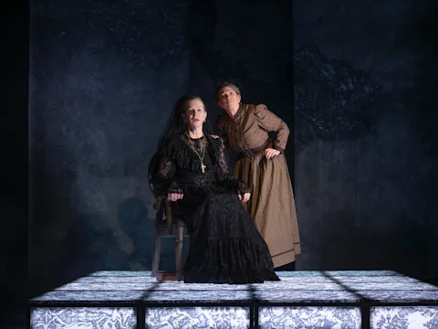 Two people in period costumes, one sitting on a chair in a black dress and the other standing in a brown dress, on an illuminated stage with a dark background.