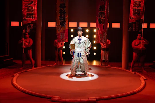 Person in decorative attire stands in a sumo ring under spotlights, surrounded by sumo wrestlers and banners.