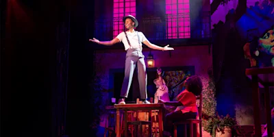 A performer stands on a table with arms outstretched on a colorful stage, while two people sit below and dramatic lighting highlights the scene.