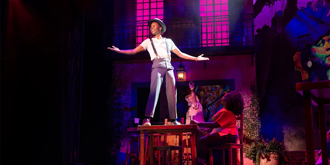 A performer stands on a table with arms outstretched on a colorful stage, while two people sit below and dramatic lighting highlights the scene.