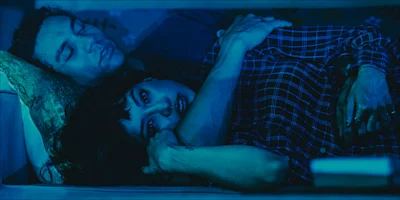 A couple lies in bed under blue lighting; one person is awake and looking forward while the other sleeps with an arm draped around them.