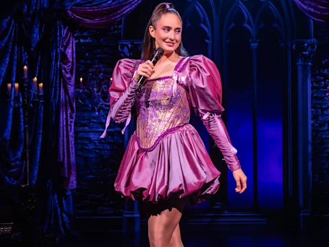 A performer in a shiny purple dress holds a microphone on stage, standing in front of a gothic-style backdrop with stone walls and dramatic purple curtains.