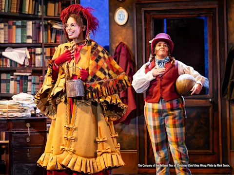 Two actors in colorful period costumes perform on stage; one holds a kettle and smiles while the other stands with hand on chest and a globe under their arm, in a room filled with books.