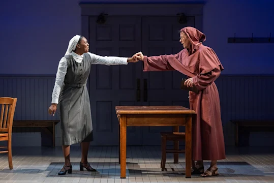 Two actors onstage, one dressed as a nun and the other as a monk, fist bump across a wooden table in a simply furnished room with gray walls and double doors.